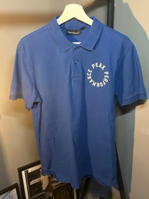 Blå Peak Performance pikétröja - Snygg blå pikétröja från Peak Performance med vit logga tryckt i cirkel på bröstet. Klassisk krage och knappar framtill. Tillverkad i mjuk bomull och har korta ärmar för en avslappnad look.