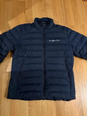 Marinblå dunjacka från Sail Racing - Snygg marinblå dunjacka från Sail Racing med quiltad design och två dragkedjeförsedda fickor framtill. Jackan har hög krage och diskret logga på bröstet. Perfekt för kalla dagar och enkel att matcha med olika outfits. Nypris 3000kr