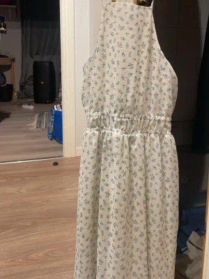 Vit blommig långklänning - Söt vit långklänning med små blå blommor och smock i midjan. Klänningen har ärmlös design och faller fint med lätt volang i kjolen. Perfekt för sommarens alla tillfällen.
