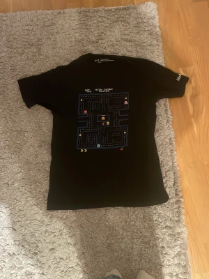 Svart Pac-Man t-shirt från Uniqlo - Cool svart t-shirt från Uniqlo x MoMA med klassiskt Pac-Man-motiv på bröstet. Perfekt för dig som gillar retrospel och vill sticka ut med en unik design. Skön passform.