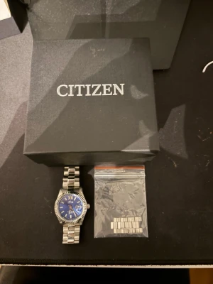 Citizen klocka med blå urtavla - Stilren Citizen klocka i rostfritt stål med blå urtavla och romerska siffror. Länkarmband i metall och räfflad boett. Levereras med originalask och extra länkbitar. Pris kan diskuteras.