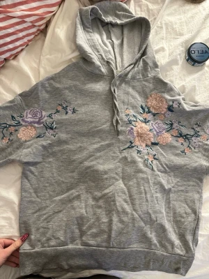 Grå hoodie med broderade blommor - Grå hoodie med huva och dragsko, dekorerad med broderade blommor i lila och ljusrosa på ärmar och bröst. Mjuk och skön, perfekt för en chill stil. Kortärmad modell som sticker ut med sina detaljer.