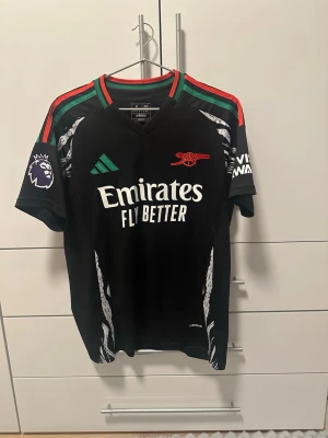 Arsenal bortatröja Adidas svart - Snygg svart Arsenal bortatröja från Adidas med gröna och röda detaljer på axlarna. Tröjan har klubbmärke, sponsorlogga och Premier League-märke på ärmen. Tillverkad i lätt och ventilerande material, perfekt för fotbollsträning eller match.