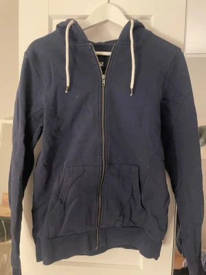 Mörkblå hoodie med dragkedja 157 - Mörkblå hoodie från 157 med vit dragsko i huvan och hel dragkedja framtill. Tröjan har två stora fickor på magen och ribbade muddar vid ärmslut och nederkant. Perfekt för en avslappnad och stilren look.