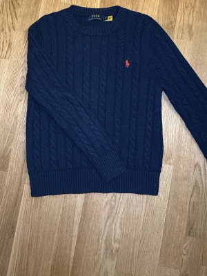 Mörkblå kabelstickad tröja Polo Ralph Lauren - Kabelstickad mörkblå tröja från Polo Ralph Lauren med rund halsringning och röd broderad logga på bröstet. Tröjan har långa ärmar och ribbstickade muddar vid ärmslut och nederkant. Perfekt för en stilren look. Passar som xs