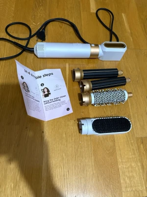 Multistyler hårverktyg med tillbehör - Multistyler för håret i vitt och guld med flera utbytbara tillbehör: två svarta locktänger, två runda borstar och en fönmunstycke. Perfekt för att skapa olika frisyrer och volym. Elektrisk med sladd och enkel att använda.