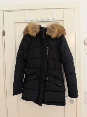 Svart dunjacka med äkta päls  - Snygg svart dunjacka med stor huva och fluffig päls i bruna toner. Jackan har långa ärmar, dragkedja framtill, två stora fickor med lock och en extra bröstficka med dragkedja. Perfekt för kalla vinterdagar. 