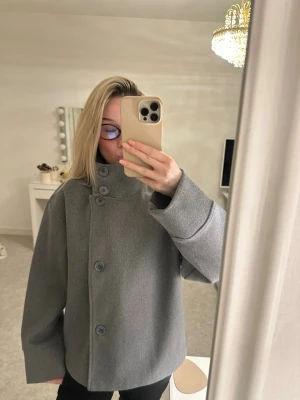 Grå kappa med stora knappar - Stilren grå kappa med hög krage och stora knappar framtill. Modellen är rak och har långa ärmar. Perfekt till höst och vår!