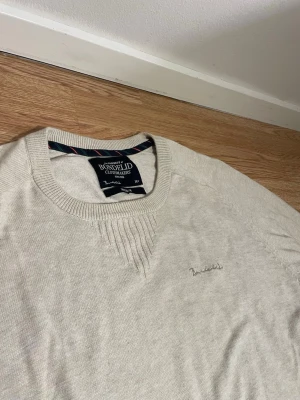 Beige tröja från Bondelid - Snygg beige tröja från Bondelid med rund halsringning och diskret broderad logga på bröstet. Tröjan har ribbade muddar vid ärmslut och nederkant samt ett stilrent mönster vid halsen. Perfekt för en clean och avslappnad look.