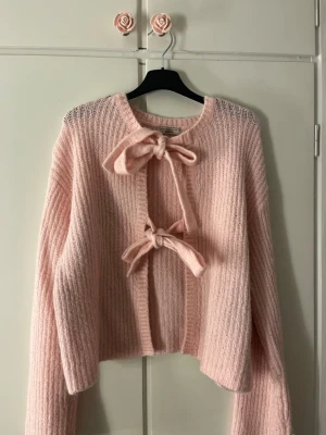 Ljusrosa stickad cardigan med rosetter - Supersöt ljusrosa stickad cardigan med två stora rosetter framtill. Tröjan har ribbad struktur, lång ärm och croppad passform. Perfekt för dig som gillar gulliga detaljer och mjuka material.