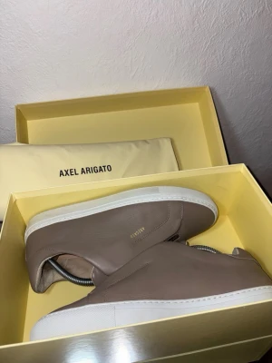 Beige sneakers från Axel Arigato - Stilrena beige sneakers från Axel Arigato i skinn med vit platt sula och silverfärgad dragkedja framtill. Minimalistisk design utan snörning och diskret guldtext på sidan. Perfekta för dig som gillar clean och modern look. Använ max 1 gång, storlek 42, kartong och dustbag medföljer! Kan kännas lite tajta. 