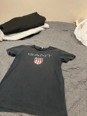 Svart GANT t-shirt med logga - Svart t-shirt från GANT med klassisk logga och sköldtryck framtill. T-shirten har rund hals och korta ärmar, perfekt för en stilren och avslappnad look. Tillverkad i mjuk bomull som känns skön mot huden.