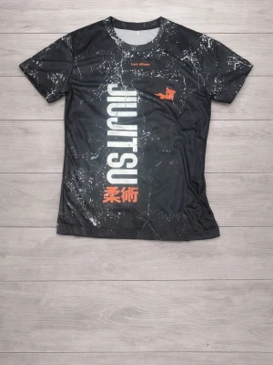 Svart Jiu Jitsu t-shirt Team Ishimaki - Snygg svart t-shirt från Team Ishimaki med vitt och orange tryck. Stort vertikalt 'JIU JITSU' tryck på framsidan, marmorerat mönster och japanska tecken. Tillverkad i ett lätt och stretchigt funktionsmaterial, perfekt för träning.