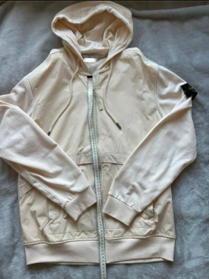 Beige hoodie med dragkedja - Snygg beige hoodie med dragkedja framtill och justerbar huva. Hoodien har långa ärmar, ribbade muddar och fickor på sidorna. Materialet ser ut att vara mjukt bomullstyg, perfekt för en avslappnad stil. Liten svart detalj på ärmen. Köpt på Vinted, osäker på om den är äkta därav priset!