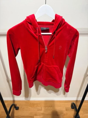 Röd velour hoodie från Juicy Couture - Snygg röd hoodie i mjuk velour från Juicy Couture. Klassisk zip-up modell med huva, fickor framtill och broderad logga på bröstet. Perfekt för en chill och trendig look. Dragsko i huvan och ribbade muddar vid ärmar och nederkant.