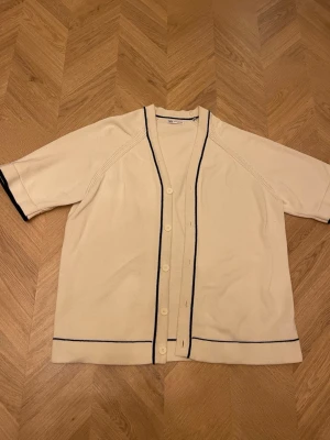 Beige kofta med svarta detaljer Zara - Kortärmad beige kofta från Zara med svarta tunna ränder längs knäppningen, ärmsluten och nederkanten. Koftan har v-ringning och knäpps med ljusa knappar framtill. Perfekt för en stilren och enkel look.