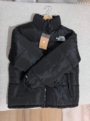 Svart pufferjacka från The North Face - Snygg svart pufferjacka från The North Face med hög krage och dragkedja framtill. Jackan har logga på bröstet och ryggen, samt två sidofickor. Perfekt för kalla dagar och riktigt trendig stil.