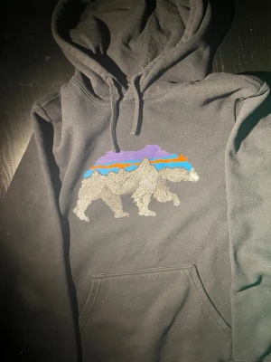 Svart hoodie med björntryck - Säljer en svart sällsynt hoodie med stor huva och magficka. På bröstet finns ett stort tryck av en björn med färgglada berg i lila, blått och orange. Hoodien har dragsko i huvan och är gjord i mjukt bomullsmaterial. Perfekt för dig som gillar streetwear och coola prints. Hoodien är i mycket bra skicka och utan deffekter🙌