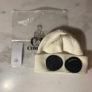  C.P,CompanyMössa - Trendiga vit stickad mössa med ikoniska goggles framtill – perfekt för streetwear fits och vinterlooks. Mjuk och skön passform som sitter bra på huvudet. Kommer med originalpåse och tag. Onesizestorlek som passar de flesta. Väldigt fint skick – redo att användas direkt.  📦 Snabb frakt efter köp 💬 Pris kan diskuteras 🔥 Paketpris vid fler köp