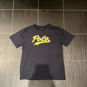 Mörkblå Polo t-shirt med gul text - Snygg mörkblå t-shirt från Polo med stor gul logga på bröstet. Klassisk rund hals och korta ärmar. Perfekt för dig som gillar enkel men stilren design. Tillverkad i mjukt material som känns skönt mot huden. DENNA TRÖJA VILL DU EJ MISSA!!!