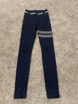 Mörkblå leggings från Stronger - Mörkblå leggings från Stronger med vita prickiga ränder på ena benet och logga i midjan. Tight passform och stretchigt material som passar perfekt till träning eller chill. Snygg och sportig look med hög midja.