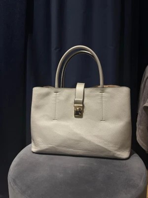 Beige handväska med gulddetalj - Stilren beige handväska i skinn med två handtag och avtagbar axelrem. Väskan har en elegant guldfärgad låsdetalj framtill och en klassisk, rektangulär form. Perfekt för dig som gillar minimalistisk design och neutrala färger.