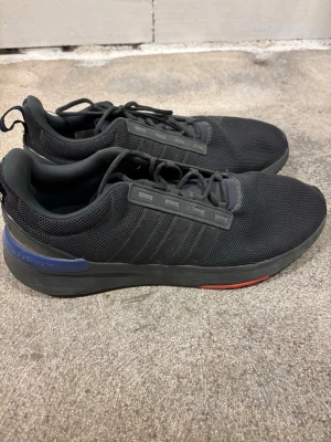 Svarta Adidas sneakers med mesh - Snygga svarta sneakers från Adidas med klassiska tre ränder på sidan. Ovandelen är i mesh för bra andningsförmåga och skon har snörning. Diskreta detaljer i blått och rött på sulan ger en cool touch. Perfekta för dig som gillar stilrena och sportiga skor.