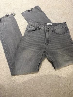 Grå jeans med råa benslut - Säljer ett par grå jeans med raka ben och råa, fransiga benslut. Klassisk femficksmodell i mjuk bomullsdenim. Perfekta för en avslappnad och trendig look.