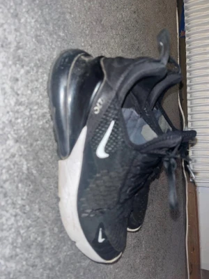 Svarta Nike Air Max 270 sneakers - Säljer ett par svarta Nike Air Max 270 sneakers med vit sula och tydlig Air-bubbla i hälen. Skorna har mesh-överdel för bra ventilation, klassisk Nike-logga på sidan och snörning framtill. Perfekta för dig som gillar sportig och modern stil.