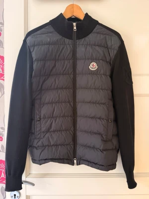 Svart dunjacka från Moncler - Snygg svart dunjacka från Moncler med quiltad front och ärmar i stickad ull. Jackan har dragkedja framtill, ribbad krage och Moncler-logga på bröstet. Perfekt för dig som vill ha en stilren och bekväm jacka med exklusiv känsla. Den är köpt på som herr modell men funkar perfekt som dam jacka också. Priset går absolut att diskutera bara att höra av sig 