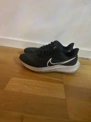 Svarta Nike sneakers med vit sula - Snygga svarta sneakers från Nike med vit sula och stor vit swoosh på sidan. Skorna har snörning och en sportig, låg profil. Ovandelen är i syntetmaterial som andas och sulan är platt och bekväm för träning eller vardagsbruk.