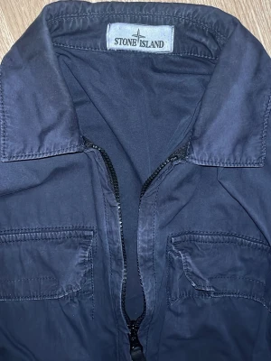 Mörkblå overshirt från Stone Island - Snygg mörkblå overshirt från Stone Island med klassisk patch på ärmen och två bröstfickor. Jackan har dragkedja framtill och är tillverkad i ett lätt bomullsmaterial. Perfekt för vår/sommar.