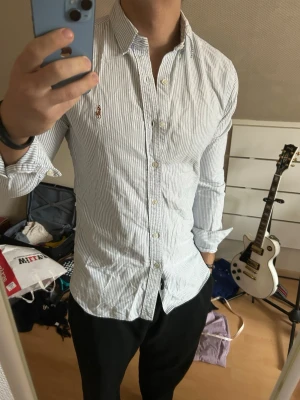 Randig skjorta från Ralph Lauren - Snygg vit skjorta med tunna blå ränder från Ralph Lauren. Klassisk krage, knäppning framtill och långärmad modell. Ikoniska broderade loggan på bröstet. Perfekt för en stilren look.