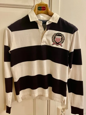 Randig rugbytröja från Gant - Säljer en långärmad rugbytröja från Gant med breda svarta och vita ränder. Tröjan har krage, knäppning vid halsen och ett broderat märke med sköld och lagerkrans på bröstet. Perfekt för en sportig och avslappnad stil. Storlek 164cm