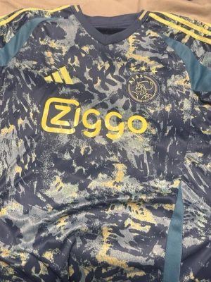 Ajax tränings t-shirt Adidas blå/gul - Ajax tränings t-shirt från Adidas i blått med gult och ljusblått mönster. Tröjan har Ajax klubbmärke och Ziggo-logga på bröstet samt tre gula ränder på axlarna. Tillverkad i funktionsmaterial som passar perfekt till träning.
