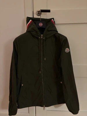 Moncler Windbreaker - Stilren mörkgrön vindjacka från Moncler i modellen Moncler Grimpeurs. Jackan har Moncler-logga på ärmen och snygga rödvita detaljer på huvan. NFC funkar! Storlek 2.