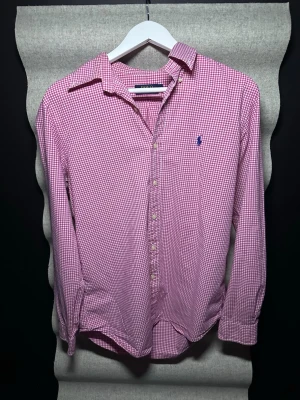 Rutig rosa skjorta från Polo Ralph Lauren - Klassisk långärmad skjorta från Polo Ralph Lauren i rosa och vitt rutigt mönster. Skjortan har knappar framtill, krage och den ikoniska blå loggan broderad på bröstet. Tillverkad i mjuk bomull, perfekt för en avslappnad men stilsäker look.