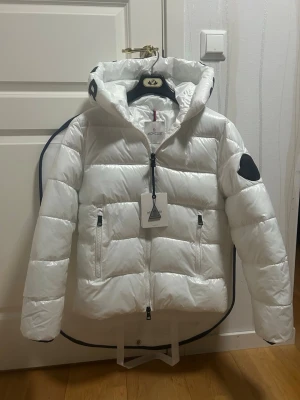 Vit dunjacka från Moncler med huva - Snygg vit dunjacka från Moncler med quiltad design och stor huva. Jackan har svarta detaljer, dragkedja framtill och två praktiska fickor med dragkedja. Perfekt för kalla dagar och riktigt stilren look.