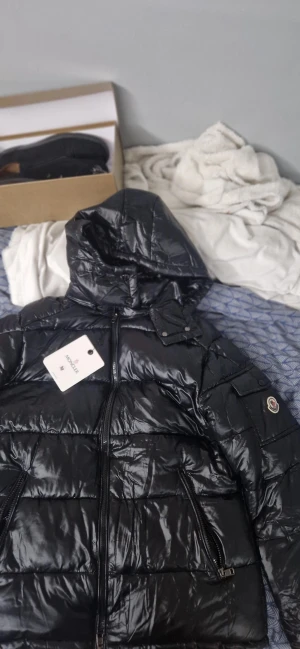 Svart pufferjacka från Moncler - (Helt ny och oanvänd)Säljer en svart pufferjacka från Moncler med klassisk logotyp på bröstet och coolt serietryck på insidan. Jackan har blanka ytor, blå och röda detaljer och knappar med Moncler-gravyr. Perfekt för dig som vill ha en snygg och varm jacka med exklusiv känsla.