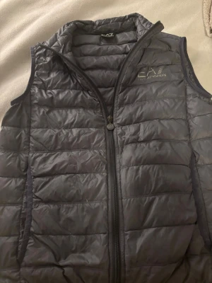 Mörkgrå dunväst från EA7 Armani - Snygg mörkgrå dunväst från EA7 Emporio Armani med quiltad design och dragkedja framtill. Västen har hög krage och diskret logga på bröstet. Perfekt för lager på lager och ger en sportig vibe till din outfit.