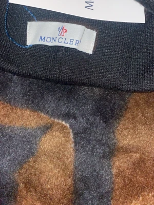 Svart Moncler mössa med ränder - Snygg svart mössa från Moncler med klassiska blå och röda ränder samt Moncler-logga framtill. Tillverkad i mjukt stickat material som sitter skönt på huvudet. Perfekt accessoar för dig som vill ha en stilren och trendig look.