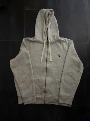 Grå zip hoodie från Polo Ralph Lauren - Grå hoodie med dragkedja från Polo Ralph Lauren. Klassisk broderad logga på bröstet, huva med snören och två stora fickor framtill. Mjuk bomullsblandning och ribbade muddar för extra komfort. Storlek XL passar L