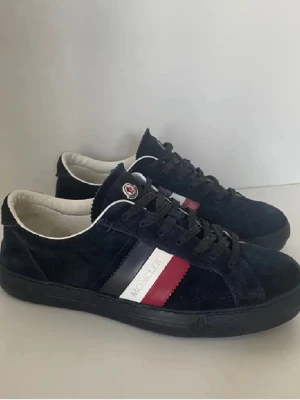 Moncler sneakers i mocka  - Snygga sneakers från Moncler i mörkblå mocka med svarta snören och klassiska Moncler-ränder i vitt, rött och blått på sidan. Låg modell med rund tå och vit insida. Perfekta för dig som gillar stilrena och exklusiva sneakers.