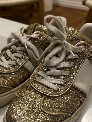 Valentino glittriga sneakers guld/vit - Snygga Valentino Garavani sneakers med glittrig guldfinish och vita detaljer. Skorna har vita snören, rund tå och platt sula. Märkeslogga på plösen och klassisk siluett som sticker ut. Perfekta för att sätta lite extra stil på din outfit. Ganska så använda men välanvända, där av priset!❤️(Enkelt att bara bytta snörerna). Inga nitar saknas! Äkthetsbevis finns!