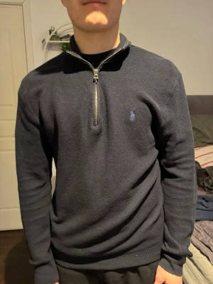 Mörkblå halvzip från Ralph Lauren - Mörkblå stickad tröja från Ralph Lauren med halv dragkedja och broderad logga på bröstet. Tröjan har ribbade muddar vid ärmslut och nederkant samt hög krage. Perfekt för en stilren och avslappnad look. Bra skick, sparsamt använd. Stor i storlek 