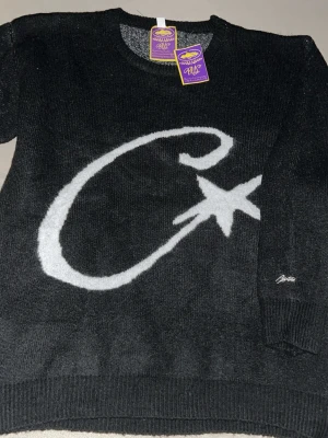 Svart stickad tröja från corteiz (Baggy) - Cool svart stickad tröja från corteiz med ett stort vitt logo på framsidan. Tröjan har rund halsringning och ribbade muddar. Perfekt för dig som gillar grafiska tryck och vill ha något unikt i garderoben. 