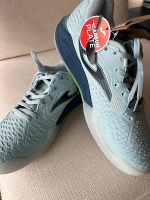 Ljusblå padel/ tennisskor från Joma - Snygga ljusblå padelskor från Joma med grå och mörkblå detaljer. Skorna har en robust sula med carbon plate för extra stabilitet och grepp. Helt oanvända med lappen kvar på skorna. Först till kvarn!