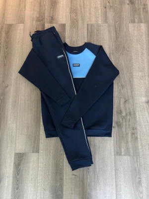 Mörkblå loungewear-set från MZKENZIE - Snyggt loungewear-set från MZKENZIE i mörkblått med ljusblå detalj på tröjan. Setet består av sweatshirt och matchande byxor med resår i midjan. Perfekt för chill dagar hemma eller när du vill vara bekväm men ändå se fräsch ut. Tar emot bud. Storleken är ca 160-165,lite baggy.