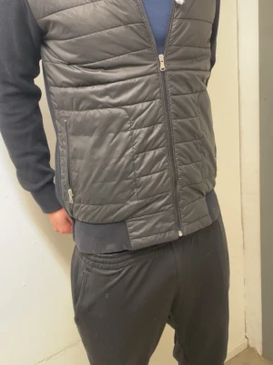 Svart cardigan - Stilren svart overshirt med quiltad framsida och mjuka stickade ärmar och rygg. Dragkedja framtill och två sidofickor med dragkedja. Perfekt lager-på-lager-plagg för en avslappnad look. Materialmix av polyester och bomull.