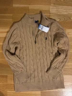 Beige kabelstickad tröja Polo Ralph Lauren - Kabelstickad beige tröja från Polo Ralph Lauren med halv dragkedja och hög krage. Tröjan har ribbade muddar och nederkant samt den klassiska mörkblå logotypen broderad på bröstet. Perfekt för lager på lager och en stilren look.